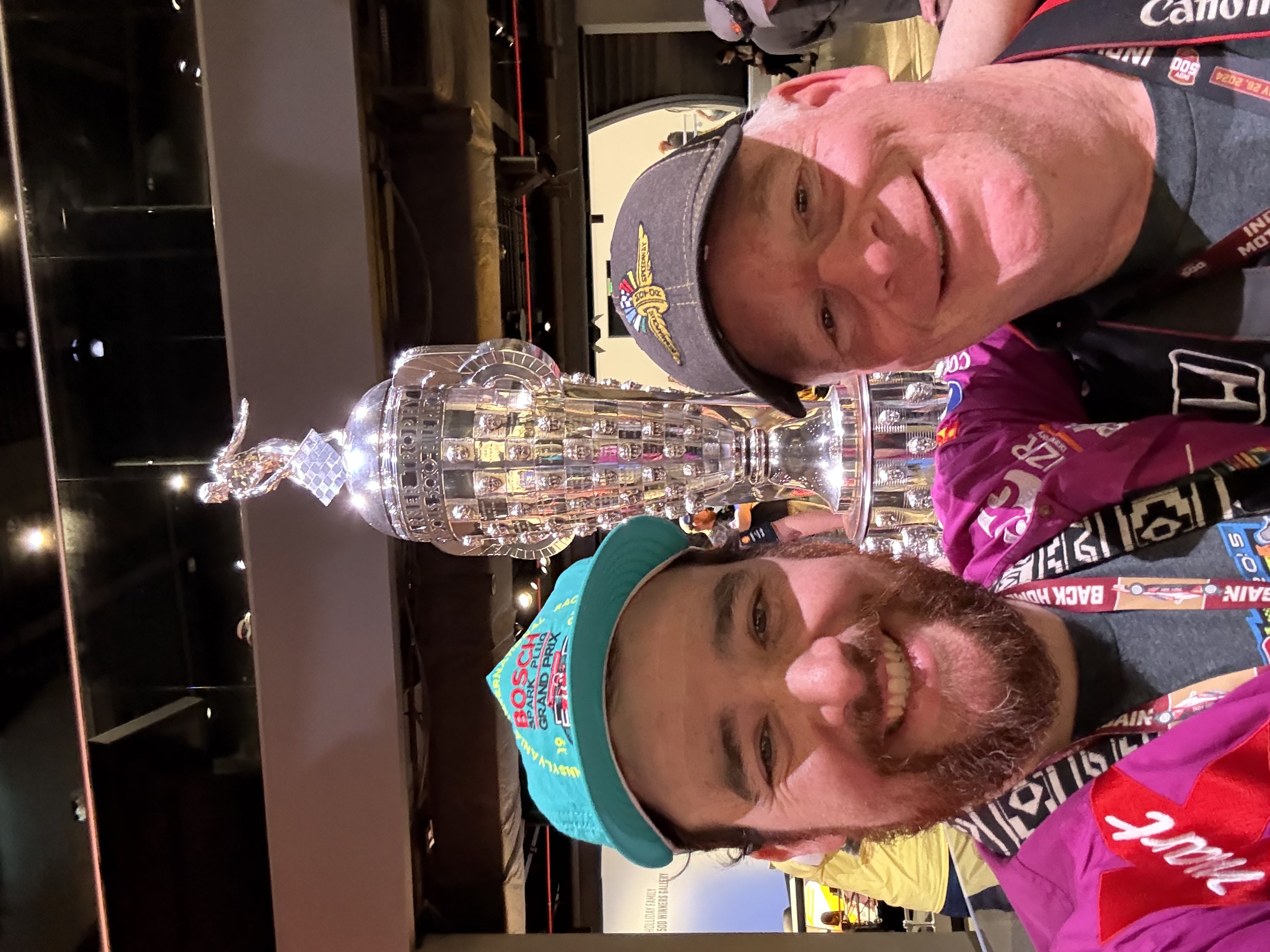 Father and son in colorful racing gear smiling for a selfie in front of the Borg Warner Trophy at Indianapolis Motor Speedway